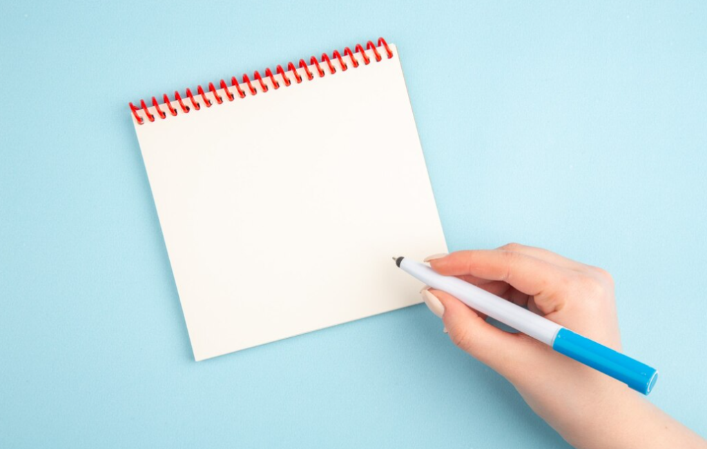 hand holds a pen near the opened notebook in the blue background
